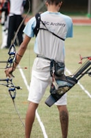 A person wearing an archery uniform stands on a grassy field holding a bow. The shirt displays the words 'CSC Archery Team.' A quiver full of arrows is attached to their waist, and they display a tattoo on their right calf. The field is marked with white lines, and there are other archers visible in the background.
