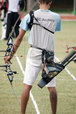 A person wearing an archery uniform stands on a grassy field holding a bow. The shirt displays the words 'CSC Archery Team.' A quiver full of arrows is attached to their waist, and they display a tattoo on their right calf. The field is marked with white lines, and there are other archers visible in the background.