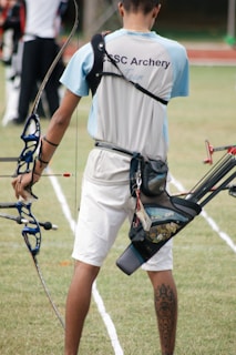 A person wearing an archery uniform stands on a grassy field holding a bow. The shirt displays the words 'CSC Archery Team.' A quiver full of arrows is attached to their waist, and they display a tattoo on their right calf. The field is marked with white lines, and there are other archers visible in the background.