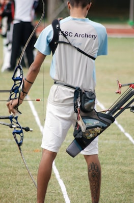 A person wearing an archery uniform stands on a grassy field holding a bow. The shirt displays the words 'CSC Archery Team.' A quiver full of arrows is attached to their waist, and they display a tattoo on their right calf. The field is marked with white lines, and there are other archers visible in the background.