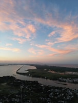 A landscape view featuring a river dividing a land area with scattered residential buildings near the bottom, open green spaces, and a horizon that stretches to the ocean. Above, the sky displays scattered clouds with warm hues of orange and pink, creating a serene atmosphere.
