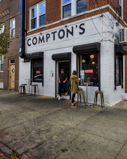 A street corner coffee shop with a brick façade and large sign reading 'Compton's.' Two people are standing outside near the entrance, with stools and a small table set up on the sidewalk. The building features a white and brown color scheme, creating a cozy, neighborhood feel.