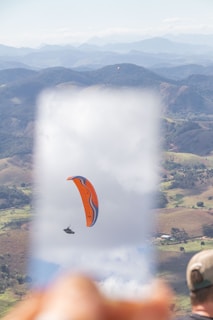 A paraglider with an orange and blue canopy soars through the sky above a mountainous landscape. The scene is framed by a blurred foreground, possibly showing a hand holding a camera or a transparent device. The background consists of rolling hills and a clear sky, creating a sense of freedom and adventure.