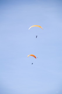 Two paragliders with bright orange sails float against a clear blue sky. The image captures them in mid-air, showcasing the sport of paragliding.