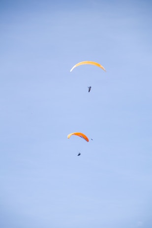 Two paragliders with bright orange sails float against a clear blue sky. The image captures them in mid-air, showcasing the sport of paragliding.