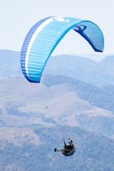 A paraglider with a large blue canopy soars above a vast mountainous landscape. The pilot is suspended below, holding onto the lines and wearing a helmet.