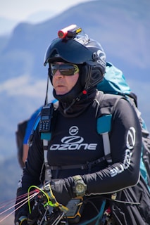 A person is wearing a helmet with a camera mounted on top, sunglasses, and a jacket labeled with 'Ozone'. They are equipped with various gear including a radio and a wristwatch, and appear ready for an outdoor adventure, possibly paragliding. The background features a blurred mountainous landscape.