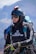 A person is wearing a helmet with a camera mounted on top, sunglasses, and a jacket labeled with 'Ozone'. They are equipped with various gear including a radio and a wristwatch, and appear ready for an outdoor adventure, possibly paragliding. The background features a blurred mountainous landscape.