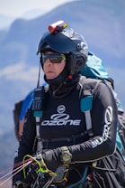 A person is wearing a helmet with a camera mounted on top, sunglasses, and a jacket labeled with 'Ozone'. They are equipped with various gear including a radio and a wristwatch, and appear ready for an outdoor adventure, possibly paragliding. The background features a blurred mountainous landscape.