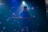 A musician is seated at a Nord Stage 3 keyboard, playing under a spotlight on a dark stage. The background is filled with dim, abstract purple lights, giving a moody ambiance.