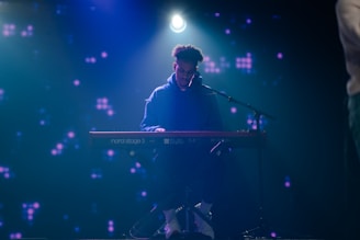A musician is seated at a Nord Stage 3 keyboard, playing under a spotlight on a dark stage. The background is filled with dim, abstract purple lights, giving a moody ambiance.