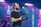 A person wearing a black t-shirt and a red and beige cap is holding a professional video camera. The background features colorful stage lights, creating a vibrant atmosphere suggestive of a live event or concert. The person has tattoos on their arm and is focused on filming.