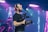A person wearing a black t-shirt and a red and beige cap is holding a professional video camera. The background features colorful stage lights, creating a vibrant atmosphere suggestive of a live event or concert. The person has tattoos on their arm and is focused on filming.