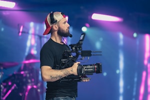 A person wearing a black t-shirt and a red and beige cap is holding a professional video camera. The background features colorful stage lights, creating a vibrant atmosphere suggestive of a live event or concert. The person has tattoos on their arm and is focused on filming.