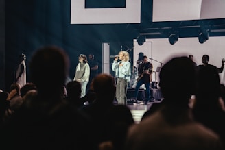 A live performance with a female singer holding a microphone on stage, accompanied by musicians playing instruments. The stage is illuminated with spotlights, and a crowd of people is visible in the foreground, watching the performance.