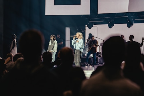 A live performance with a female singer holding a microphone on stage, accompanied by musicians playing instruments. The stage is illuminated with spotlights, and a crowd of people is visible in the foreground, watching the performance.
