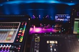 A stage is lit with vibrant colored lights, featuring musicians performing with various instruments under blue overhead lighting. In the foreground, there are electronic soundboards and music equipment. A large screen displays video content above the stage.
