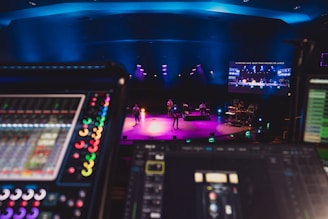 A stage is lit with vibrant colored lights, featuring musicians performing with various instruments under blue overhead lighting. In the foreground, there are electronic soundboards and music equipment. A large screen displays video content above the stage.