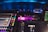 A stage is lit with vibrant colored lights, featuring musicians performing with various instruments under blue overhead lighting. In the foreground, there are electronic soundboards and music equipment. A large screen displays video content above the stage.
