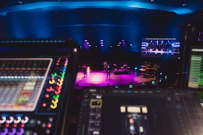 A stage is lit with vibrant colored lights, featuring musicians performing with various instruments under blue overhead lighting. In the foreground, there are electronic soundboards and music equipment. A large screen displays video content above the stage.