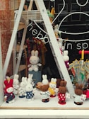 A store window display showcasing a variety of crochet bunny dolls in different colors and outfits. The bunnies are arranged in rows on shelves, with some in the front dressed in colorful polka-dotted clothes. The background includes umbrellas and decorative items, with reflections on the glass adding depth.