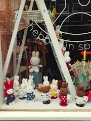A store window display showcasing a variety of crochet bunny dolls in different colors and outfits. The bunnies are arranged in rows on shelves, with some in the front dressed in colorful polka-dotted clothes. The background includes umbrellas and decorative items, with reflections on the glass adding depth.