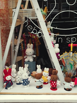 A store window display showcasing a variety of crochet bunny dolls in different colors and outfits. The bunnies are arranged in rows on shelves, with some in the front dressed in colorful polka-dotted clothes. The background includes umbrellas and decorative items, with reflections on the glass adding depth.