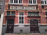 The image features an old brick building with a historic sign reading 'St. Antonius Meisjesschool' dated 1895. The building has decorative elements, including arched wooden doors and grid-patterned windows. The facade is adorned with ornate brickwork.