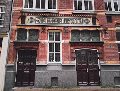 The image features an old brick building with a historic sign reading 'St. Antonius Meisjesschool' dated 1895. The building has decorative elements, including arched wooden doors and grid-patterned windows. The facade is adorned with ornate brickwork.
