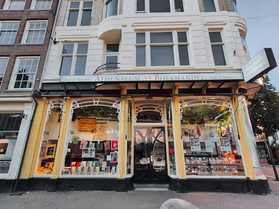 A quaint bookstore with large front windows displaying a variety of books and magazines. The building features classic architecture with decorative arches and pillars. Signage above the door and windows indicates the bookstore's name, and there are posters and promotional materials visible in the windows. The exterior color scheme includes cream and white tones, and the sidewalk in front adds to the urban setting.