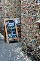 A narrow alleyway with walls completely covered in chewed gum of various colors, creating a mosaic-like texture. A wooden easel stands on the left side with a blackboard advertising a live improv comedy show tonight at 8 pm, adorned with colorful chalk drawings. A QR code is also displayed on a small sign attached to the easel.