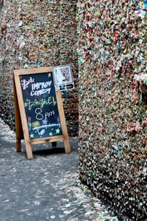 A narrow alleyway with walls completely covered in chewed gum of various colors, creating a mosaic-like texture. A wooden easel stands on the left side with a blackboard advertising a live improv comedy show tonight at 8 pm, adorned with colorful chalk drawings. A QR code is also displayed on a small sign attached to the easel.