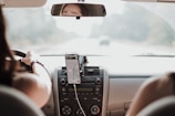 A person is driving a car with a smartphone mounted on the dashboard displaying a navigation app.
