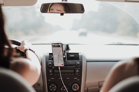 A person is driving a car with a smartphone mounted on the dashboard displaying a navigation app.