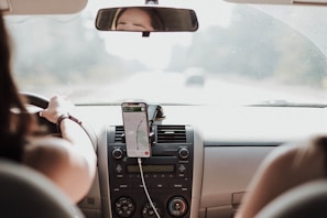 A person is driving a car with a smartphone mounted on the dashboard displaying a navigation app.