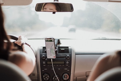 A person is driving a car with a smartphone mounted on the dashboard displaying a navigation app.