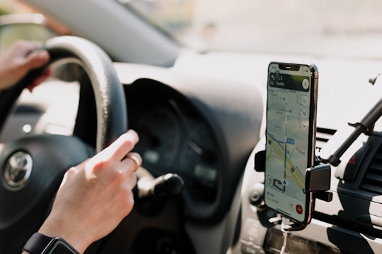 A person is driving a car with a hand on the steering wheel. A smartphone is mounted on the dashboard displaying a navigation map. The interior of the car, including the steering wheel and dashboard layout, is visible. Sunlight enters through the window, casting shadows.