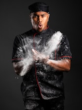 A chef wearing a black uniform with red piping claps both hands, creating a cloud of flour around him. The dramatic lighting highlights the dynamic movement and expression of focus and determination on his face. He wears a black chef's hat and stands against a plain dark background.