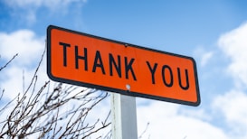An orange rectangular sign with the words 'THANK YOU' written in bold black letters. The sign is mounted on a metal pole against a backdrop of blue sky with scattered clouds and some bare tree branches.