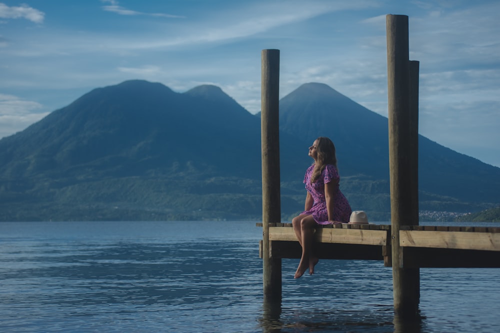a woman sitting on a dock with mountains in the background
