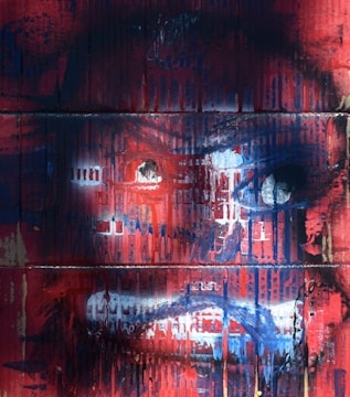 A vivid, abstract painting with a dominant face appearing in a mix of red, blue, and white brush strokes. The expression is intense, with eyes that seem to be staring forward amidst a chaotic background.