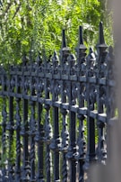An ornate, black wrought iron fence is shown with intricate spiral and spearhead designs. Lush green foliage serves as the backdrop, with sunlight filtering through the leaves, creating a play of light and shadow.