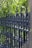 An ornate, black wrought iron fence is shown with intricate spiral and spearhead designs. Lush green foliage serves as the backdrop, with sunlight filtering through the leaves, creating a play of light and shadow.