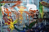 An abstract painting with a chaotic mix of colors and textures. Bold splashes of orange, green, and yellow are layered over a backdrop of muted blues and grays. The composition includes swirling patterns, dripping paint, and overlapping brush strokes.