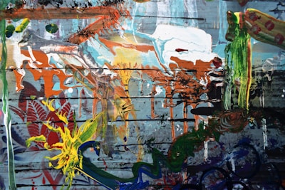 An abstract painting with a chaotic mix of colors and textures. Bold splashes of orange, green, and yellow are layered over a backdrop of muted blues and grays. The composition includes swirling patterns, dripping paint, and overlapping brush strokes.