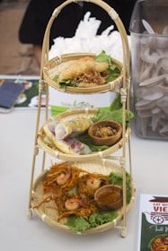 A tiered bamboo stand holds various Vietnamese dishes. The top shelf displays a fried dish on a bed of lettuce, garnished with crispy onion. The middle shelf contains fresh spring rolls and a small wooden bowl filled with a condiment. The bottom shelf holds shrimp-topped crispy noodles with a bowl of sauce.