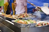 A food stall displaying trays of assorted grilled skewers and rolls. A person is seen holding skewers and tending to the food. The background shows a blue tarp and some yellow decorations.