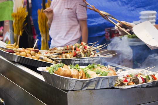 A food stall displaying trays of assorted grilled skewers and rolls. A person is seen holding skewers and tending to the food. The background shows a blue tarp and some yellow decorations.