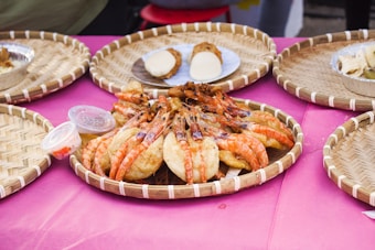 A display of fried shrimp arranged on a woven basket tray, surrounded by other assorted foods on similar trays. These sit on a pink surface, with small containers of dipping sauces nearby.