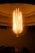 An illuminated vintage-style light bulb with visible filaments casts a warm, amber glow. The bulb is housed within a lamp, and its intricate filament structure is prominently displayed. The surrounding atmosphere is dimly lit, enhancing the bulb's brightness.